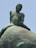 A close-up view of a weathered bronze sculpture depicting a nude male figure sitting atop a large, rounded object. The surface of the sculpture shows signs of aging, with visible patina and erosion.