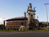 A large, historic bullring with ornate architecture stands prominently in the background, featuring arched windows and a red brick facade. In front of the bullring, there is an elaborate stone monument adorned with statues and intricate carvings. The sky is clear with a few clouds, and the scene includes a grassy area and a sidewalk in the foreground.