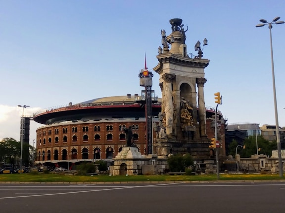 A large, historic bullring with ornate architecture stands prominently in the background, featuring arched windows and a red brick facade. In front of the bullring, there is an elaborate stone monument adorned with statues and intricate carvings. The sky is clear with a few clouds, and the scene includes a grassy area and a sidewalk in the foreground.