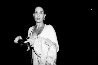 A person in a white outfit stands against a dark background. The individual holds a coat and a clutch purse, with a chain handbag strap visible. The light source illuminates the face and upper body, emphasizing the contrast with the dark surroundings.