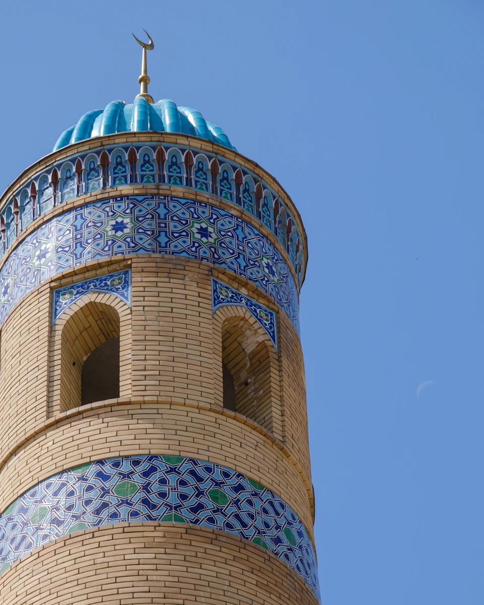 The image shows a tall, ornate brick tower with a blue dome and intricate patterns adorning its surface. The tower is situated against a clear blue sky, creating a striking contrast between the man-made structure and the natural environment. The perspective of the photo is from below, looking up at the tower, emphasizing its height and grandeur.