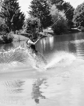 A person engaged in wakeboarding on a calm body of water, creating splashes as they maneuver. The surrounding area is lush with tall trees and greenery reflecting off the water's surface.
