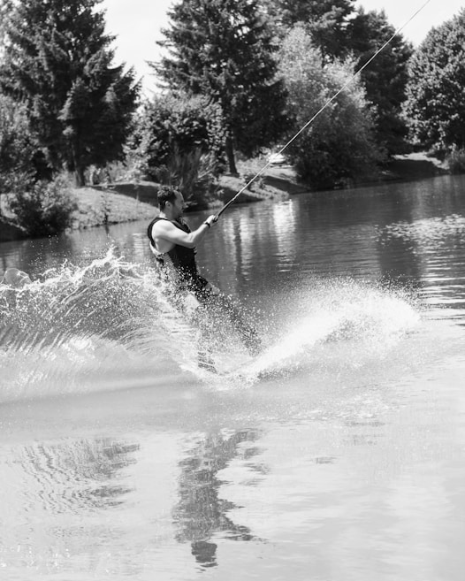 A person engaged in wakeboarding on a calm body of water, creating splashes as they maneuver. The surrounding area is lush with tall trees and greenery reflecting off the water's surface.