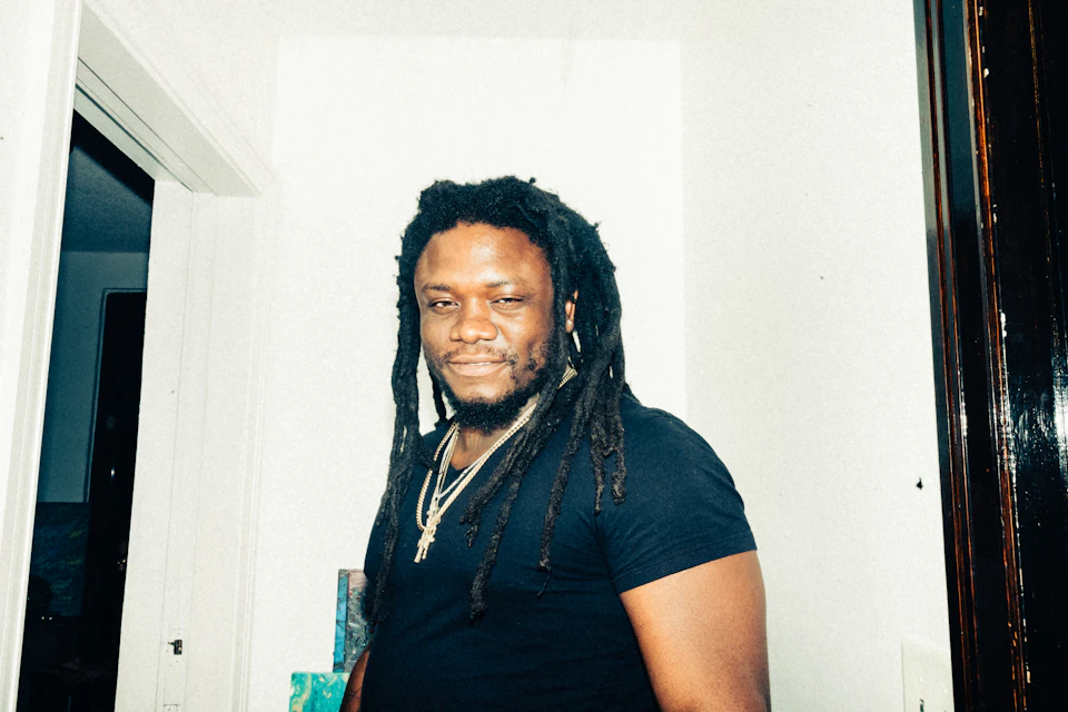 In the center of the image, a man with dreadlocks is standing against a white wall. He's dressed casually in a black t-shirt and has his arms crossed over his chest. The man's gaze is directed straight at the camera, giving off an air of confidence and self-assuredness.

The room he's in appears to be a bathroom or a bedroom, as suggested by the presence of a door on one side of the image. There are also two books visible in the background, adding to the overall atmosphere of the space.