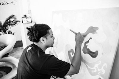 A person with braided hair is focused on painting a canvas with abstract patterns. The person is holding a paintbrush and appears to be in the process of creating art. There are some indoor plants and modern furniture in the background, including a chair and a frame on the wall.