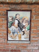 A ceramic tile mural on a brick wall depicts a group of historical figures, dressed in traditional Renaissance attire, gathered around a table laden with food. The focal point is a bearded man in a dark robe holding a long stick, possibly a conductor's baton or a similar object, with others in colorful garments. Plates of food and an ornate golden goblet are visible on the table. There is a Spanish inscription below the image.