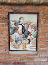 A ceramic tile mural on a brick wall depicts a group of historical figures, dressed in traditional Renaissance attire, gathered around a table laden with food. The focal point is a bearded man in a dark robe holding a long stick, possibly a conductor's baton or a similar object, with others in colorful garments. Plates of food and an ornate golden goblet are visible on the table. There is a Spanish inscription below the image.