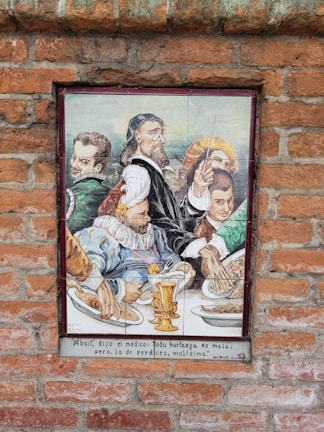 A ceramic tile mural on a brick wall depicts a group of historical figures, dressed in traditional Renaissance attire, gathered around a table laden with food. The focal point is a bearded man in a dark robe holding a long stick, possibly a conductor's baton or a similar object, with others in colorful garments. Plates of food and an ornate golden goblet are visible on the table. There is a Spanish inscription below the image.