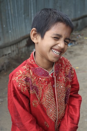 A smiling boy is wearing a red, embellished traditional outfit. He is standing outdoors near a corrugated metal wall and a dirt pathway.
