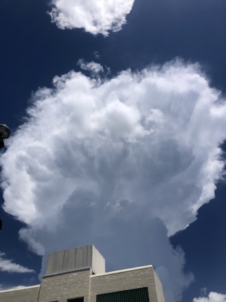 A towering cumulus cloud formation rises dramatically into the sky above a building with a flat roof. The cloud is billowy and white, contrasting sharply against the deep blue sky. Part of the building is visible, with a textured exterior wall and a metallic top.