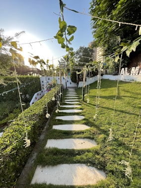 A garden pathway made of stone slabs is bordered by lush green grass and hedges. Overhead, strings with leaves and decorations hang, casting shadows on the path. In the background, there