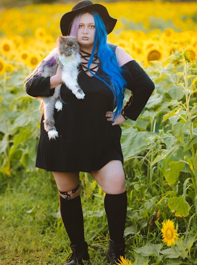 a woman with blue hair is holding a cat in a field of sunflowers
