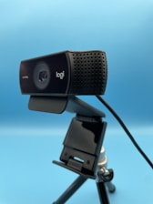 A black webcam mounted on a tripod against a blue background. The webcam has detailing with several small holes on one side, and branding on the front. A cable is visible, connected to the device.