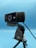 A black webcam mounted on a tripod against a blue background. The webcam has detailing with several small holes on one side, and branding on the front. A cable is visible, connected to the device.