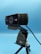 A black webcam mounted on a tripod against a blue background. The webcam has detailing with several small holes on one side, and branding on the front. A cable is visible, connected to the device.
