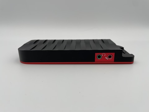 A sleek, black electronic device with a textured top surface and a red trim. On one side, there are two audio jacks with icons for headphones and a microphone.