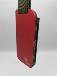 A sleek and modern red electronic device with a side view showing various ports, including USB-C and HDMI. It has a sturdy olive green strap for portability. The surface is smooth, displaying a logo and text related to video capture.