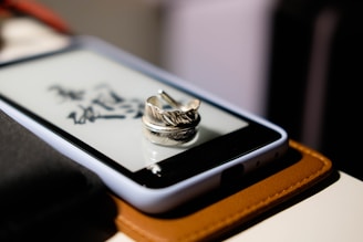 A metallic feather-shaped ring rests on the screen of a smartphone encased in a white cover. The phone is placed on a brown leather surface, and some black calligraphic text is visible on the screen beneath the ring.