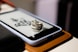A metallic feather-shaped ring rests on the screen of a smartphone encased in a white cover. The phone is placed on a brown leather surface, and some black calligraphic text is visible on the screen beneath the ring.