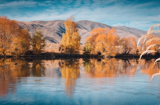 Golden-hued trees reflect peacefully on the surface of a calm lake, with rugged mountains in the background under a partially cloudy sky. The scene captures the essence of autumn with vibrant foliage and serene water.