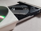 A close-up view of an open CD/DVD drive with a partially visible disc. The drive's mechanism, including the laser lens and spindle, is clearly visible. The background is a plain light-colored surface.