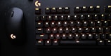 A black mechanical keyboard with illuminated keys, alongside a matching black gaming mouse with an illuminated logo. The keyboard keys are lit with a subtle red and yellow backlight, creating a contrast against the dark background.