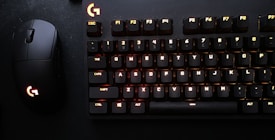 A black mechanical keyboard with illuminated keys, alongside a matching black gaming mouse with an illuminated logo. The keyboard keys are lit with a subtle red and yellow backlight, creating a contrast against the dark background.