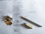 A sheet of paper with hand-drawn sketches and notes, featuring a pencil sketch of a mobile app interface design. A sharpened pencil, an eraser, and a pencil sharpener with shavings are placed on the paper. The sketches include arrows and annotations, suggesting planning or brainstorming.