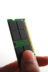 A close-up view of a person holding a green computer RAM module between their thumb and fingers. The module has black chips and a golden edge connector visible.