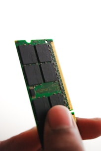 A close-up view of a person holding a green computer RAM module between their thumb and fingers. The module has black chips and a golden edge connector visible.