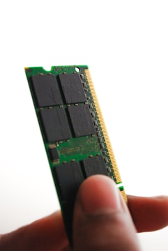 A close-up view of a person holding a green computer RAM module between their thumb and fingers. The module has black chips and a golden edge connector visible.