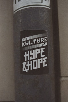 A weathered pole displays a monochromatic sticker with bold, stylized text reading 'The Kulture of Hype & Hope'. The surrounding environment suggests an urban setting with a gritty texture.