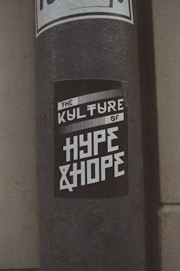 A weathered pole displays a monochromatic sticker with bold, stylized text reading 'The Kulture of Hype & Hope'. The surrounding environment suggests an urban setting with a gritty texture.