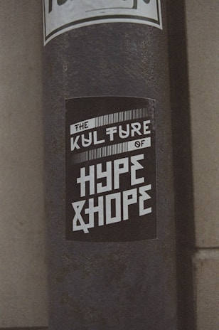 A weathered pole displays a monochromatic sticker with bold, stylized text reading 'The Kulture of Hype & Hope'. The surrounding environment suggests an urban setting with a gritty texture.