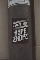 A weathered pole displays a monochromatic sticker with bold, stylized text reading 'The Kulture of Hype & Hope'. The surrounding environment suggests an urban setting with a gritty texture.