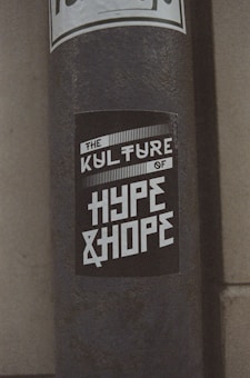 A weathered pole displays a monochromatic sticker with bold, stylized text reading 'The Kulture of Hype & Hope'. The surrounding environment suggests an urban setting with a gritty texture.