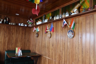 A cozy restaurant interior with wooden walls and ceiling, decorated with small international flags on the wall. Shelves above hold various ceramic jars, teapots, and figurines. A small table with a green booth seating and condiments is placed against the wall. Colorful paper decorations hang from the ceiling, and some decorative plates are mounted on the wall.