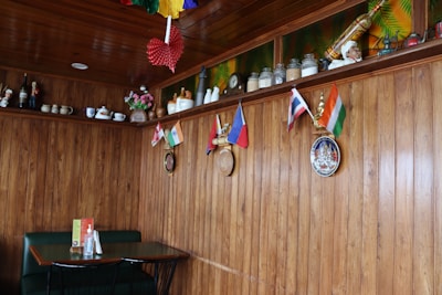 A cozy restaurant interior with wooden walls and ceiling, decorated with small international flags on the wall. Shelves above hold various ceramic jars, teapots, and figurines. A small table with a green booth seating and condiments is placed against the wall. Colorful paper decorations hang from the ceiling, and some decorative plates are mounted on the wall.