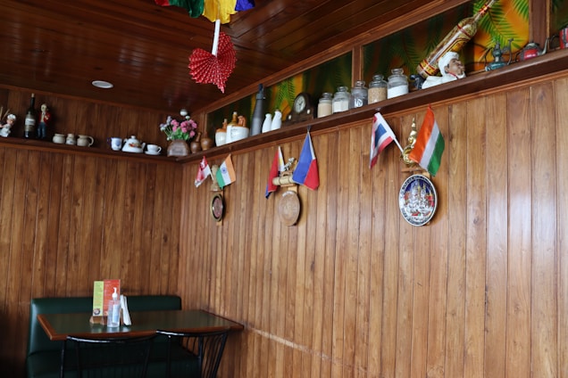 A cozy restaurant interior with wooden walls and ceiling, decorated with small international flags on the wall. Shelves above hold various ceramic jars, teapots, and figurines. A small table with a green booth seating and condiments is placed against the wall. Colorful paper decorations hang from the ceiling, and some decorative plates are mounted on the wall.