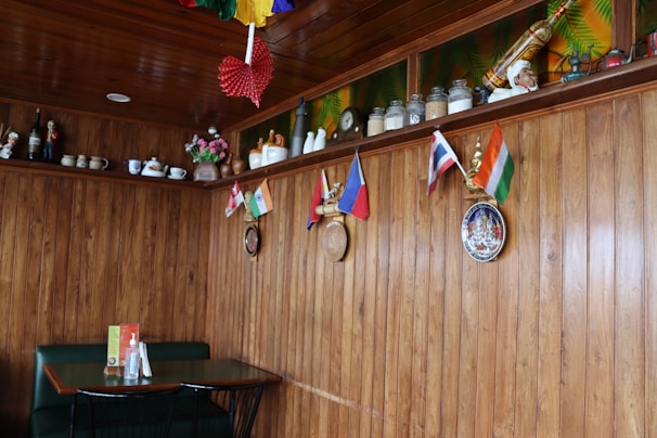 A cozy restaurant interior with wooden walls and ceiling, decorated with small international flags on the wall. Shelves above hold various ceramic jars, teapots, and figurines. A small table with a green booth seating and condiments is placed against the wall. Colorful paper decorations hang from the ceiling, and some decorative plates are mounted on the wall.