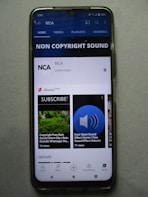 A smartphone displaying a YouTube channel named NCA, which appears to focus on non-copyright sound. There is a subscription notification bell, videos including sound effects, and a layout featuring home, videos, playlists, and channels tabs. Some of the video thumbnails and titles include 'COPYRIGHT FREE RAIN SOUND', 'DOOR OPEN SOUND EFFECT', and 'Shorts beta'. The interface includes options for home, shorts, subscriptions, and library at the bottom.