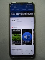 A smartphone displaying a YouTube channel named NCA, which appears to focus on non-copyright sound. There is a subscription notification bell, videos including sound effects, and a layout featuring home, videos, playlists, and channels tabs. Some of the video thumbnails and titles include 'COPYRIGHT FREE RAIN SOUND', 'DOOR OPEN SOUND EFFECT', and 'Shorts beta'. The interface includes options for home, shorts, subscriptions, and library at the bottom.