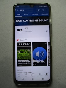 A smartphone displaying a YouTube channel named NCA, which appears to focus on non-copyright sound. There is a subscription notification bell, videos including sound effects, and a layout featuring home, videos, playlists, and channels tabs. Some of the video thumbnails and titles include 'COPYRIGHT FREE RAIN SOUND', 'DOOR OPEN SOUND EFFECT', and 'Shorts beta'. The interface includes options for home, shorts, subscriptions, and library at the bottom.