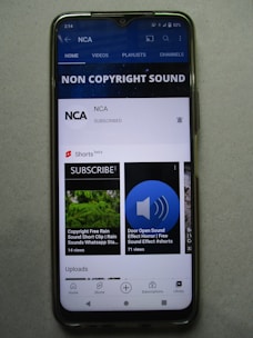 A smartphone displaying a YouTube channel named NCA, which appears to focus on non-copyright sound. There is a subscription notification bell, videos including sound effects, and a layout featuring home, videos, playlists, and channels tabs. Some of the video thumbnails and titles include 'COPYRIGHT FREE RAIN SOUND', 'DOOR OPEN SOUND EFFECT', and 'Shorts beta'. The interface includes options for home, shorts, subscriptions, and library at the bottom.