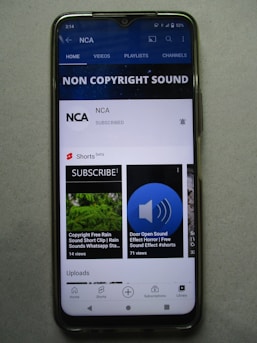 A smartphone displaying a YouTube channel named NCA, which appears to focus on non-copyright sound. There is a subscription notification bell, videos including sound effects, and a layout featuring home, videos, playlists, and channels tabs. Some of the video thumbnails and titles include 'COPYRIGHT FREE RAIN SOUND', 'DOOR OPEN SOUND EFFECT', and 'Shorts beta'. The interface includes options for home, shorts, subscriptions, and library at the bottom.