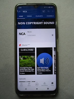 A smartphone displaying a YouTube channel named NCA, which appears to focus on non-copyright sound. There is a subscription notification bell, videos including sound effects, and a layout featuring home, videos, playlists, and channels tabs. Some of the video thumbnails and titles include 'COPYRIGHT FREE RAIN SOUND', 'DOOR OPEN SOUND EFFECT', and 'Shorts beta'. The interface includes options for home, shorts, subscriptions, and library at the bottom.