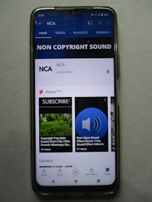A smartphone displaying a YouTube channel named NCA, which appears to focus on non-copyright sound. There is a subscription notification bell, videos including sound effects, and a layout featuring home, videos, playlists, and channels tabs. Some of the video thumbnails and titles include 'COPYRIGHT FREE RAIN SOUND', 'DOOR OPEN SOUND EFFECT', and 'Shorts beta'. The interface includes options for home, shorts, subscriptions, and library at the bottom.