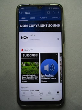 A smartphone displaying a YouTube channel named NCA, which appears to focus on non-copyright sound. There is a subscription notification bell, videos including sound effects, and a layout featuring home, videos, playlists, and channels tabs. Some of the video thumbnails and titles include 'COPYRIGHT FREE RAIN SOUND', 'DOOR OPEN SOUND EFFECT', and 'Shorts beta'. The interface includes options for home, shorts, subscriptions, and library at the bottom.