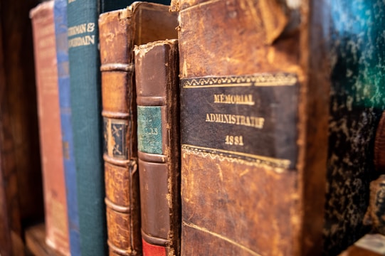A collection of old, leather-bound books with various titles visible on their spines. The books appear worn and aged, indicating they have been used frequently or are quite old. The focus is on one particular book labeled 'Memorial Administratif 1881,' suggesting a historical or administrative context.