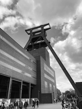 A large industrial building featuring a prominent mining headframe structure. The architecture includes geometric brick walls with long horizontal windows. People are walking near the base of the building, and the sky is partly cloudy.
